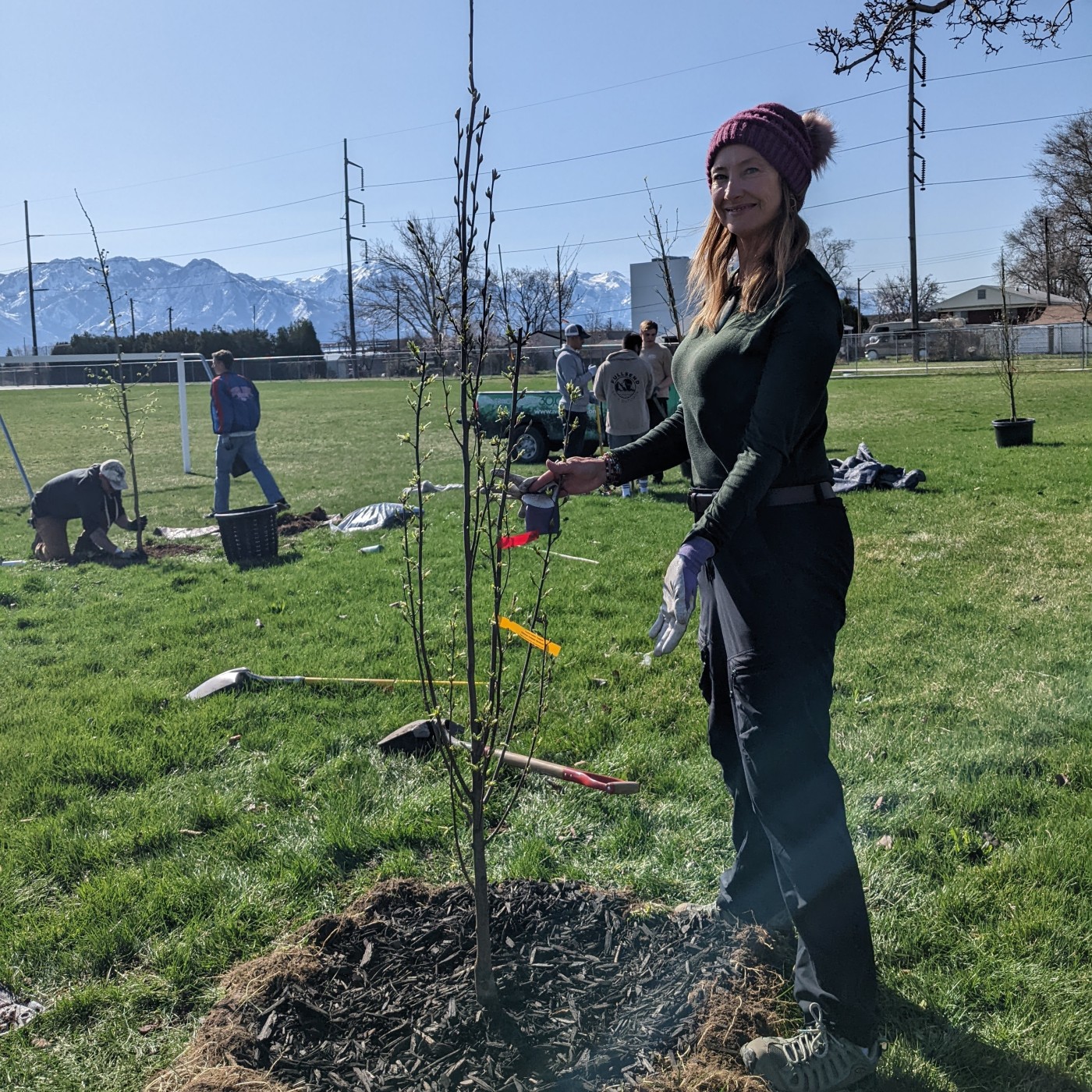 Donda holding a newly planted tree. Green grass and blue sky with mountains in the background.
