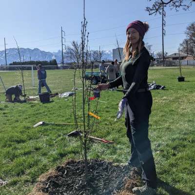 Donda holding a newly planted tree. Green grass and blue sky with mountains in the background.